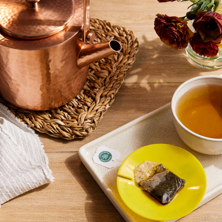 Copper kettle, Dandelion Leaf & Root Tea bag on a yellow dish, and a cup of tea promoting kidney function placed on a tray with flowers atop a wooden table. View 2
