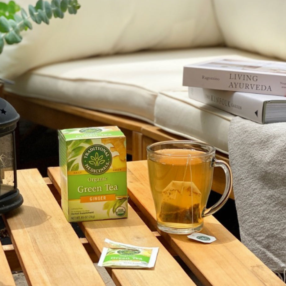 A cup of Green Tea Ginger on a wooden table with tea bags and books nearby on a cozy patio. View 2