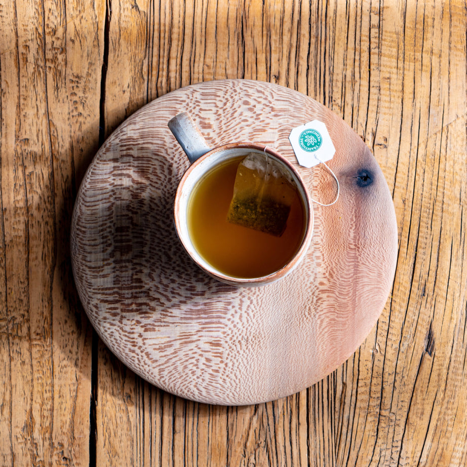 A cup of Herbal Cold Care™ Tea with a tea bag sits on a wooden coaster atop a rustic wooden table, providing soothing comfort. View 2