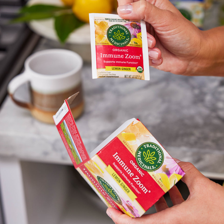 A person holding a packet of Immune Zoom® Lemon Ginger Echinacea Tea over an open box, with a mug and fresh lemon ginger in the background. View 2