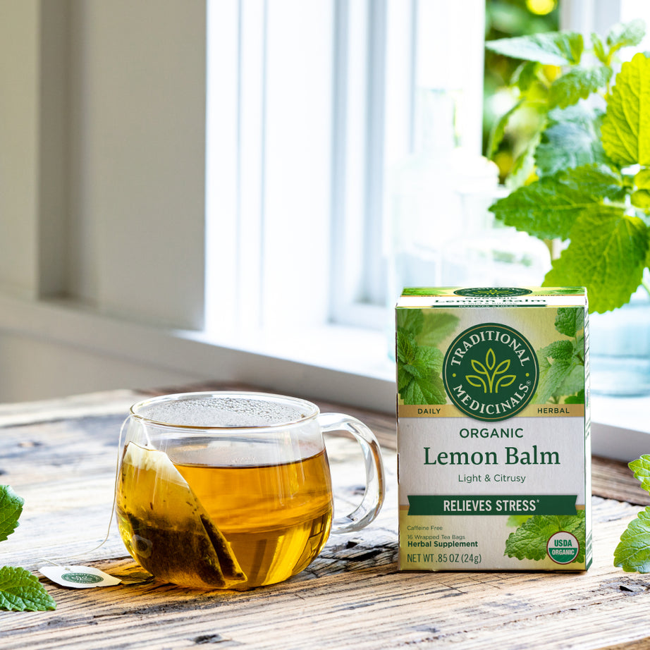 A cup of Lemon Balm Tea, renowned for its soothing effect on the nervous system, rests next to a box of Traditional Medicinals organic lemon balm on a wooden table by the window. View 2