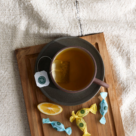 Tea cup with a lemon slice and Morning Ease® Lemon Ginger Lozenges on a wooden tray over a textured fabric background.