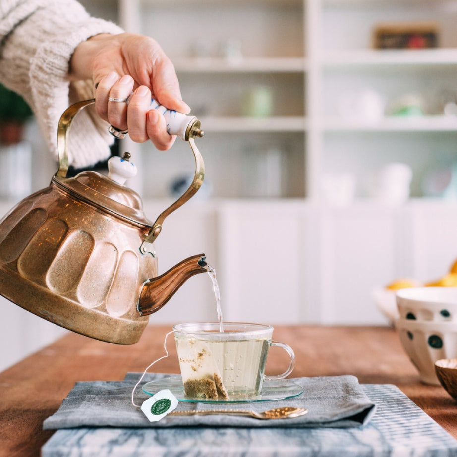 A hand carefully pours steaming hot water from a kettle into a glass cup containing a Moringa with Spearmint & Sage tea bag resting on a wooden table. View 2