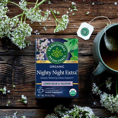 A box of Nighty Night Extra® Tea sits beside a cup, surrounded by fresh herbs and valerian root on a wooden table, inviting you to enjoy deep sleep with its soothing blend.