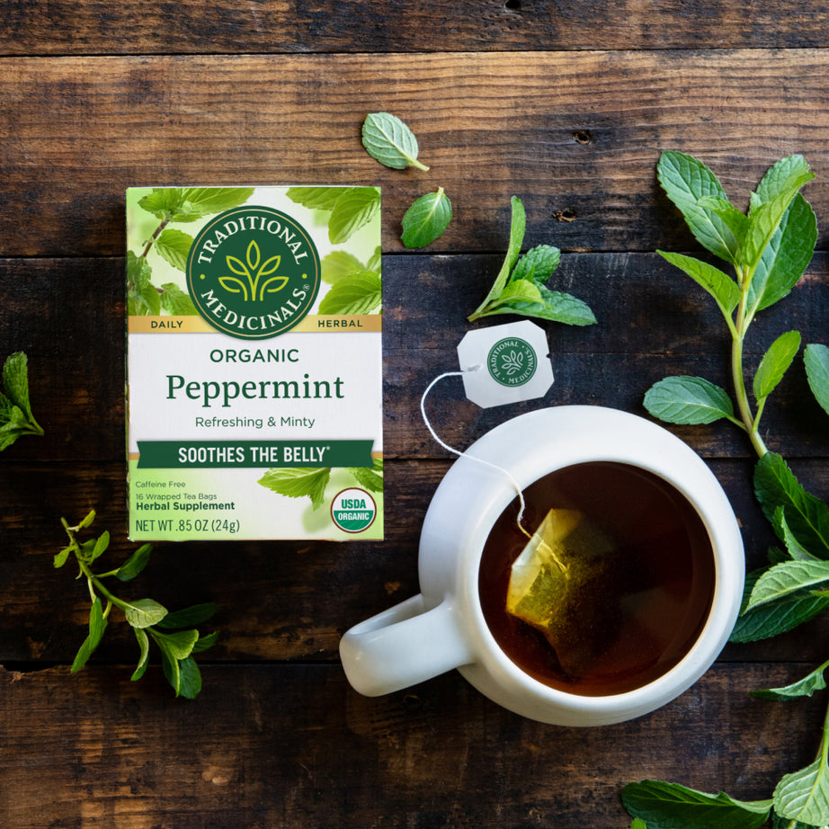 A box of Peppermint Tea sits beside a steaming cup on a wooden table, surrounded by fresh mint leaves. This soothing blend is perfect for easing digestive discomfort while providing a refreshing flavor. View 2