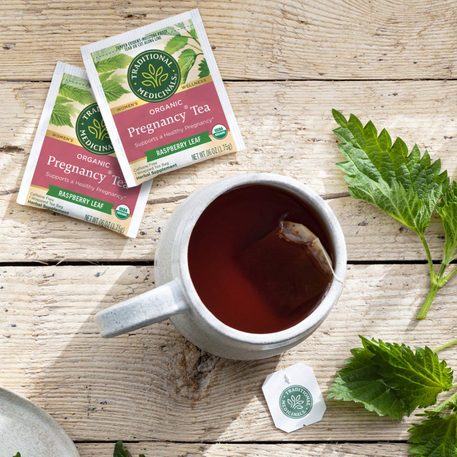 A cup of Pregnancy® Tea rests on a wooden table, infused with the promise of a healthy pregnancy. Surrounded by tea packets and green leaves, this tea offers a soothing moment for soon-to-be mothers. View 2