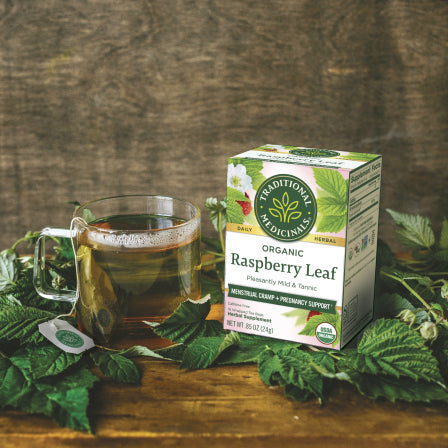 A glass cup of herbal tea sits beside a box of Raspberry Leaf Tea on a wooden table surrounded by green leaves, offering natural support for menstrual cramps. View 2