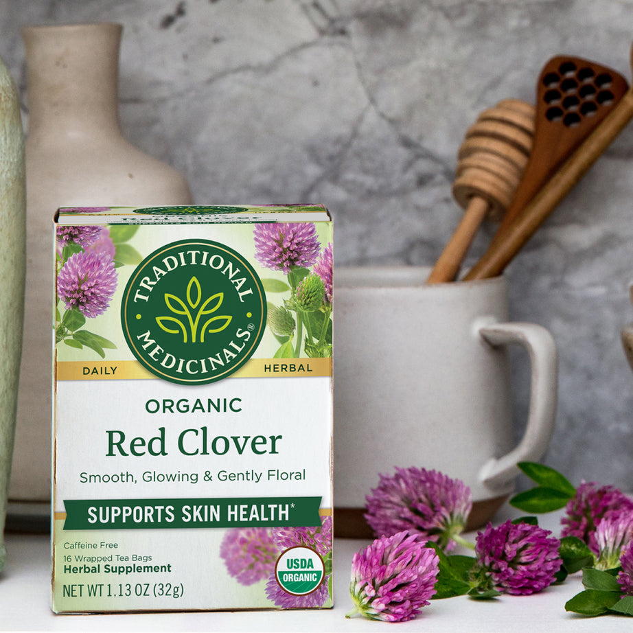 A box of Traditional Medicinals' Red Clover Tea sits surrounded by cups and honey dippers, perfect for enjoying a soothing brew that supports skin health. View 2