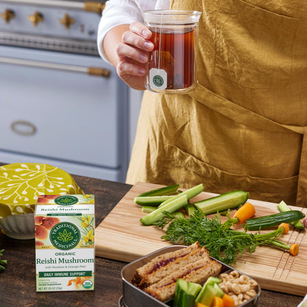 In a cozy kitchen, a person holds a steaming cup of tea, surrounded by fresh vegetables, bread, and a box of Reishi Mushroom with Rooibos & Orange Peel Tea, renowned as the