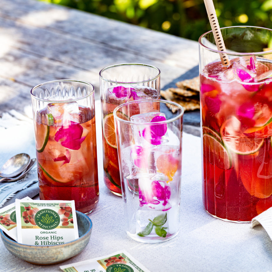 A quartet of glasses filled with refreshing iced Rose Hips & Hibiscus Tea rests on a table outside, beautifully decorated with vibrant flowers and lime slices. View 2
