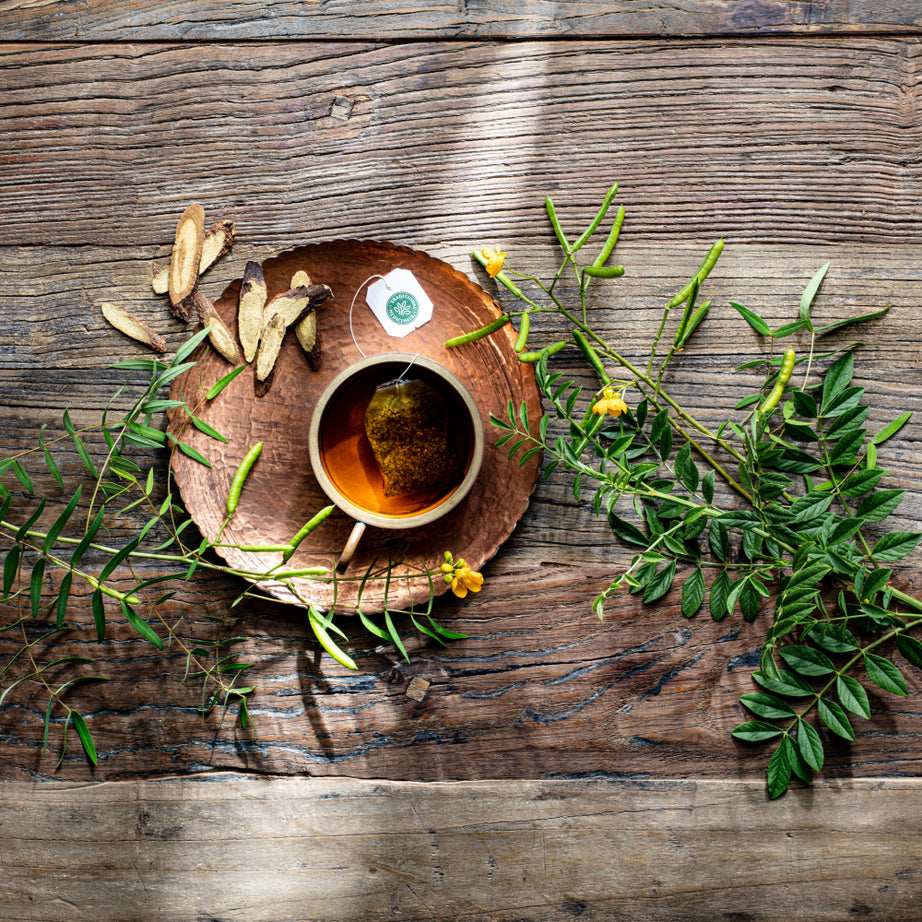 A cup of Smooth Move® Chamomile Tea on a wooden table is surrounded by green leaves and dried roots, offering gentle overnight constipation relief. View 2