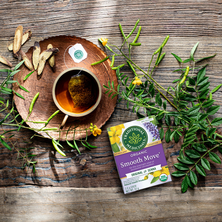 A cup of senna tea sits next to an opened box of Smooth Move® Tea on a rustic wooden table, surrounded by leaves and roots, offering gentle overnight relief. View 2