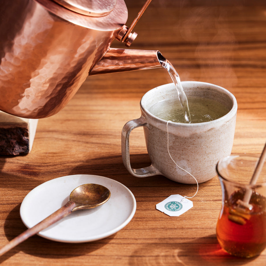 A copper kettle pours hot water into a mug containing an organic tea bag of Spearmint Tea. Next to it, honey in a jar and a spoon are placed on the wooden table, creating the perfect setup for a refreshing beverage. View 2