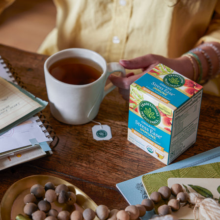 A person enjoys Stress Ease® Immune Tea infused with adaptogens at a table, alongside a box of the same stress-relief tea, notebooks, and decorative beads.