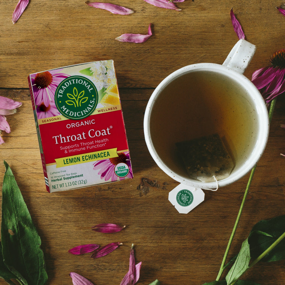 A box of Throat Coat® Lemon Echinacea Tea is placed next to a steaming mug, surrounded by flower petals on a wooden surface. Ideal for promoting throat health and supporting your immune system. View 2