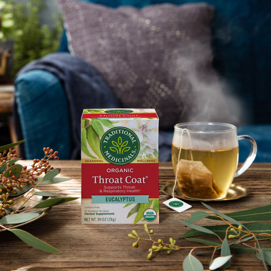 A box of Throat Coat® Eucalyptus Tea sits invitingly on a table, surrounded by eucalyptus leaves and a steaming cup, promoting throat health. A cozy couch in the background beckons for rest and relaxation, offering the perfect setting to enjoy this soothing herbal medicine. View 2