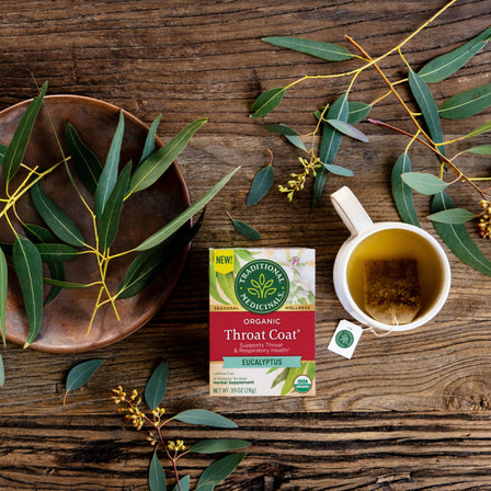 The Throat Coat® Eucalyptus Tea is beautifully displayed alongside a cup containing a tea bag and eucalyptus leaves on a wooden surface, highlighting the blend's emphasis on respiratory health.