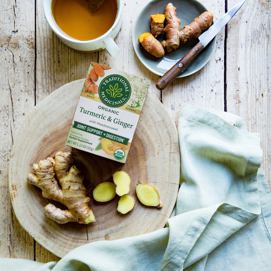 Ginger and turmeric rest on a wooden board next to a cup of meadowsweet tea and a box of Turmeric & Ginger with Meadowsweet Tea on a rustic table. View 2
