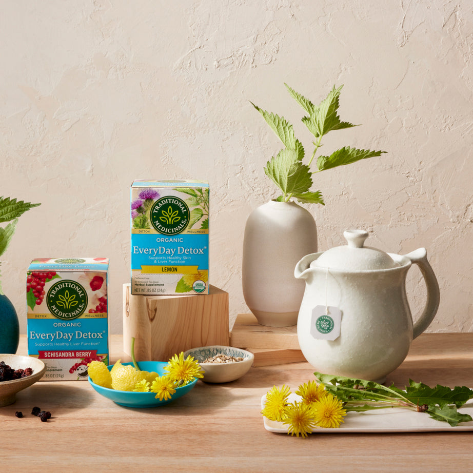 A tea setup featuring EveryDay Detox® Lemon Tea boxes from Traditional Medicinals, accompanied by a teapot, a vase with yellow flowers, and lemons, is arranged on a wooden table. It's an ideal choice for individuals looking to support liver and kidney function while promoting vibrant health and healthy skin. View 2