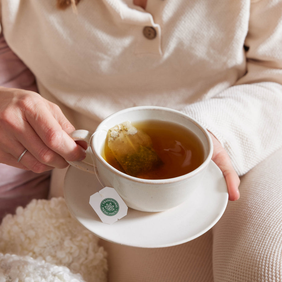 A person wearing a white shirt sits comfortably on a cozy blanket, savoring a soothing cup of Ginger & Chamomile Tea with a tea bag. View 2
