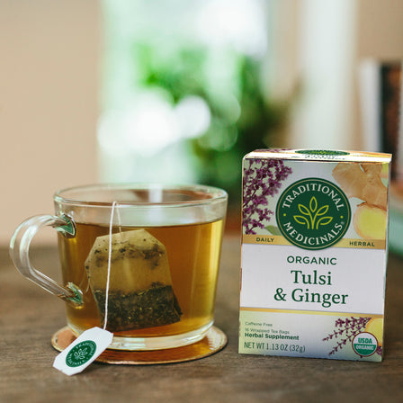 A cup of Tulsi & Ginger Tea with stress support, brewed from a teabag, sits next to a box of Traditional Medicinals on a wooden table.