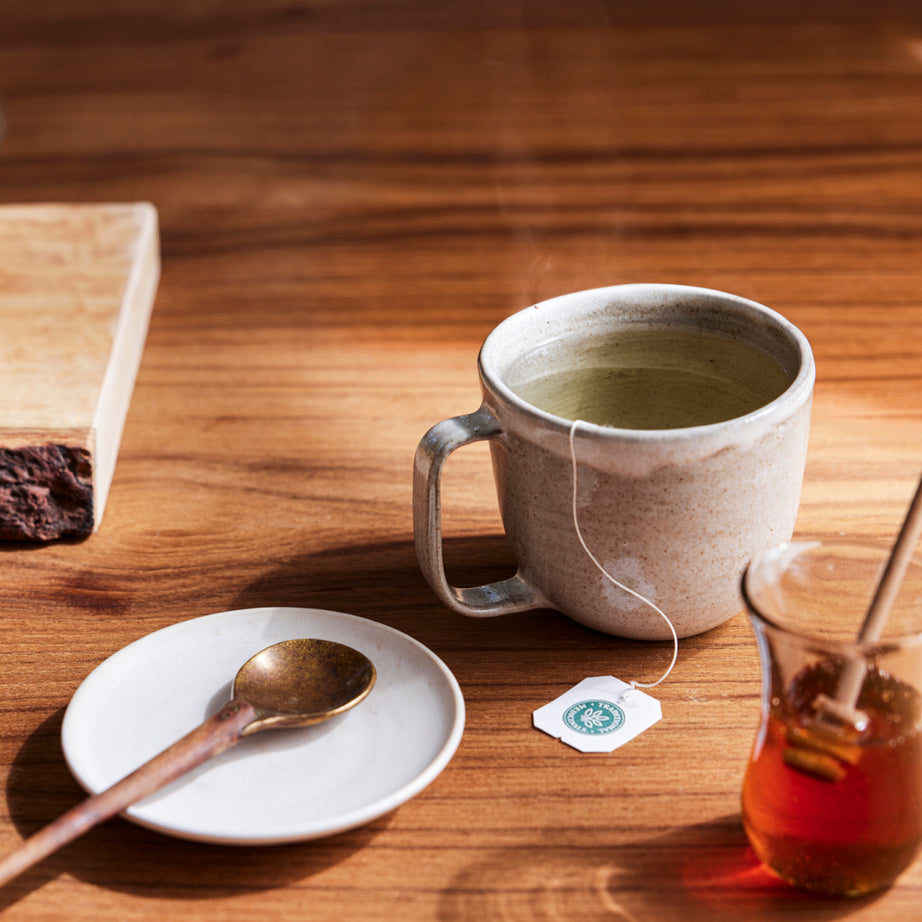 A steaming cup of Tulsi & Ginger Tea, honey jar, and spoon rest on a wooden table, offering the perfect stress support. View 2